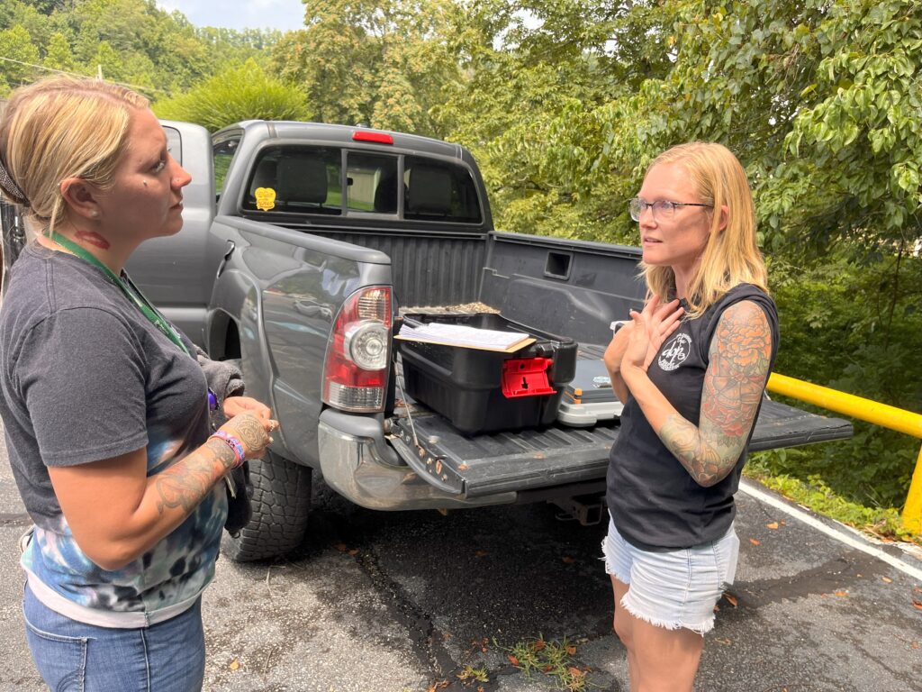 Two blonde women stand behind a truck with an open gate. The women are wearing grey and black t-shirts. One is wearing shorts and the other jeans. They are talking animatedly with their hands.