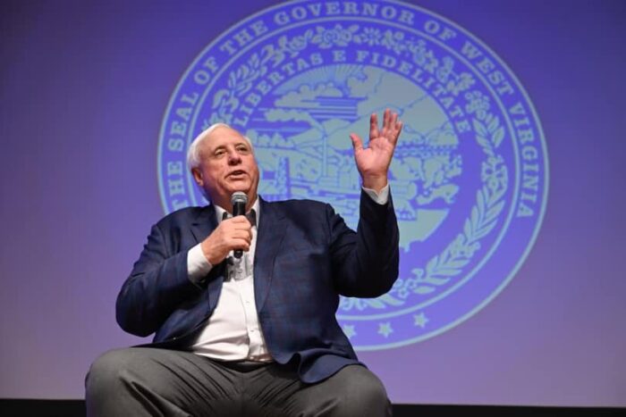 Gov. Jim Justice sits on a stool on a stage, holding a microphone. He is wearing a suit with no tie. Behind him, the governor's seal is projected onto a wall.