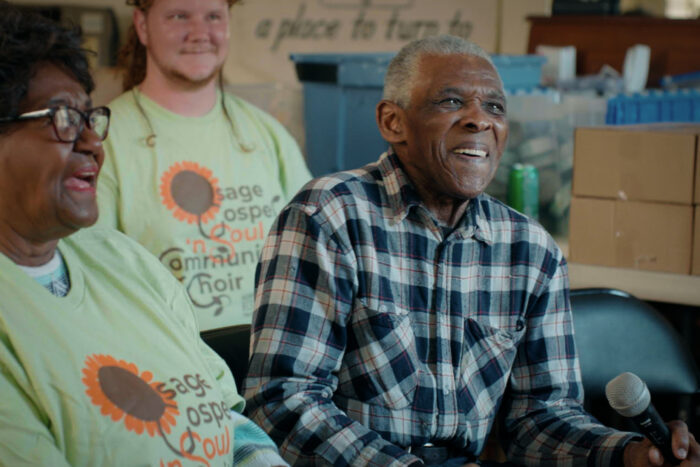 A group of happy people sing together. An older man holds a microphone and smiles.