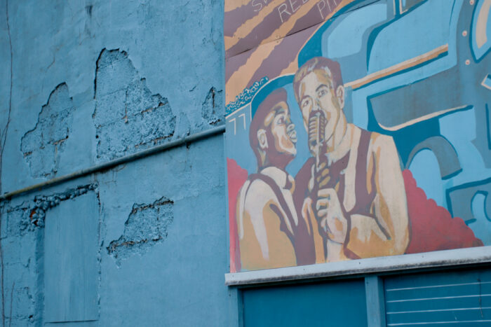 A mural of two musicians around a microphone hangs on a cracked exterior wall painted blue.