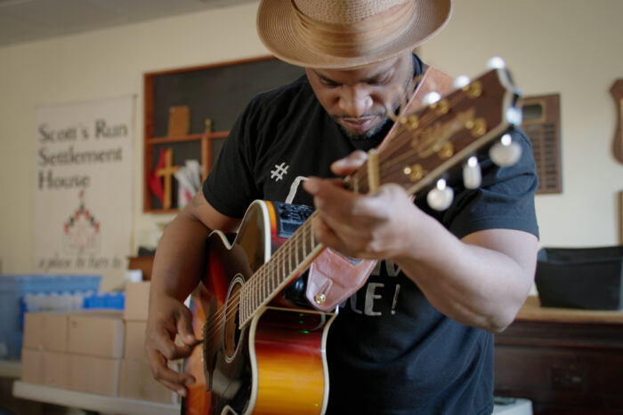 And African American man in a black t-shirt and a straw hat bows his head as he strums a guitar.