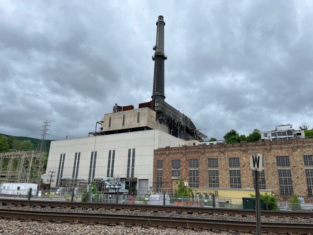 A brick building with a tall smokestack sits idle against an overcast sky with a pair of railroad tracks cutting across the foreground.