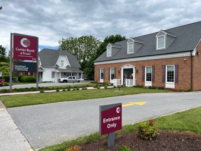 The entrance to a bank branch showing a red sign and a brick building with a gray-shingled roof on an overcast day.