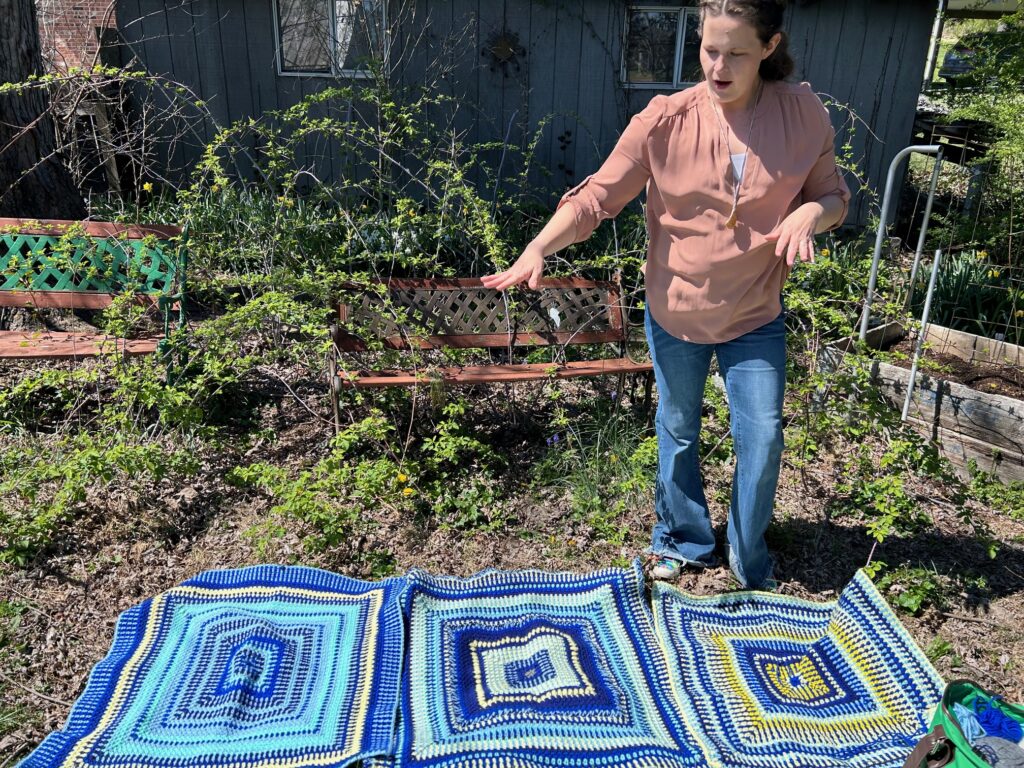 A woman displays large blue and white and blue and yellow squares of a crocheted blanket on a piece of ground outside a garden.