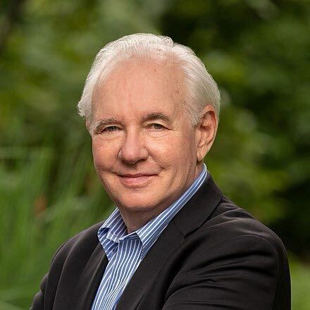 A man in a 3/4 portrait looks at camera while wearing a blue gingham shirt under a charcoal grey jacket. The man is smiling slightly with his left eyebrow slightly tilted up. Behind him a bokeh effect gives the impression of foliage and greenery.