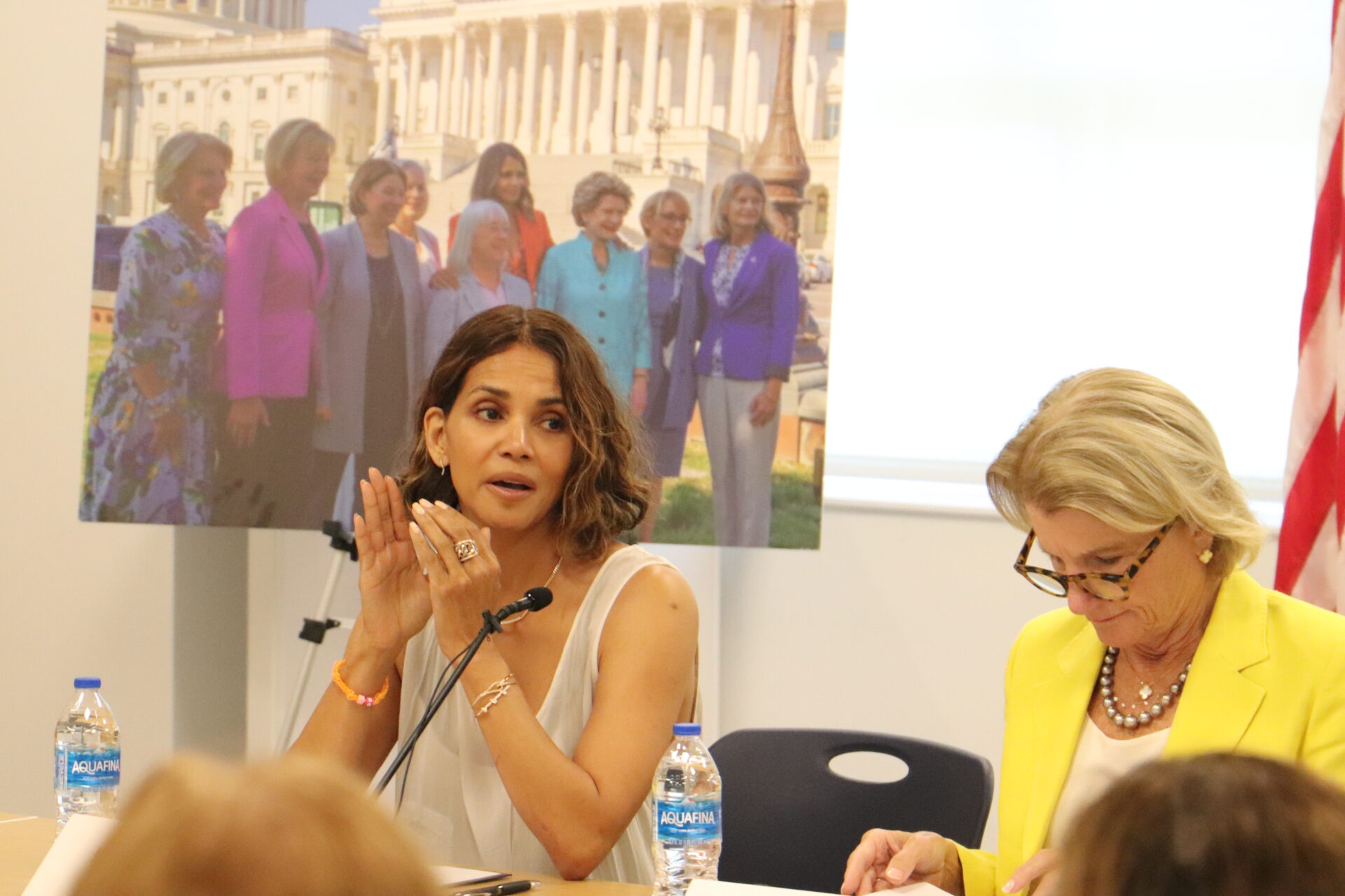 A woman wearing a sleeveless dress clasps her hands as she speaks to a group as a woman wearing a yellow jacket puts on her glasses to read a document.