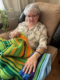 An older woman sits in tan chair looking over a crocheted blanket sitting across her lap.