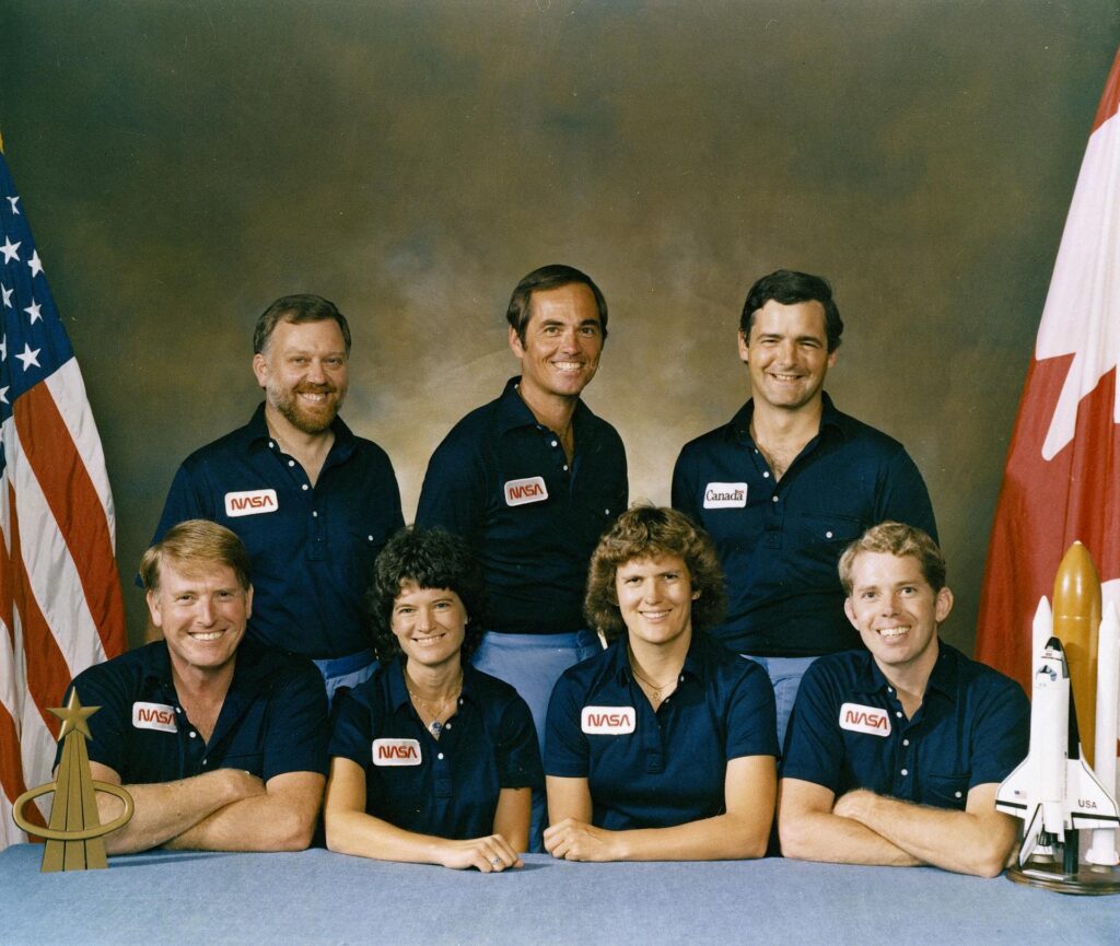 A group of seven astronauts are bracketed by the U.S. and Canadian flags in an official portrait for their space shuttle mission.