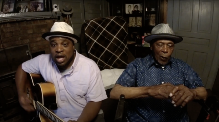 Two African American men wearing hats sit in a living room playing music.