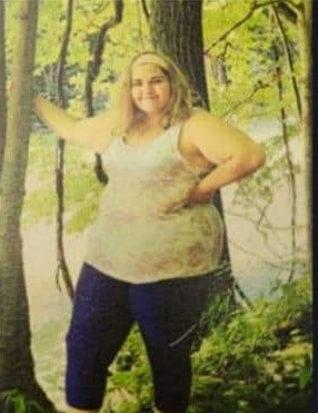 A teenage girl stands before a backdrop of a forest with her hand on her hip.