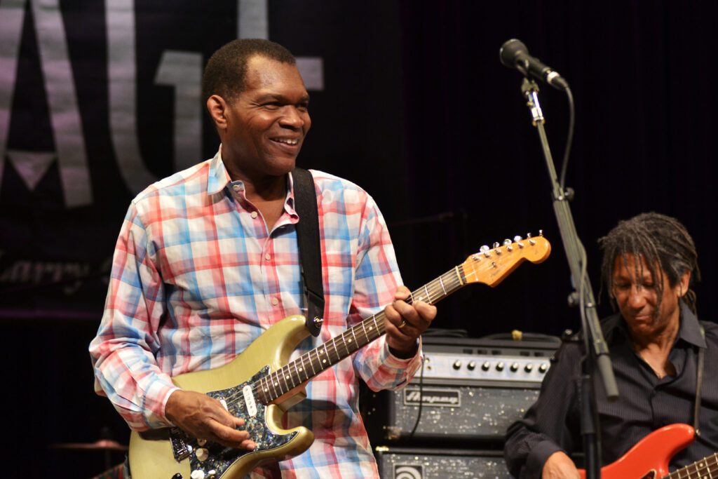 A man wearing a checkered shirt plays the guitar and smiles.