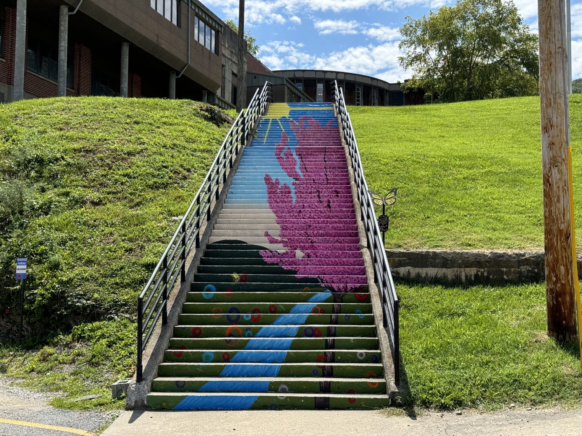 A colorful mural on a set of cement steps depicts a purple tree by a stream under a blue sky.