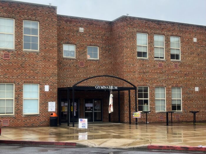 A red-brick building with white lettering atop a black awning reads "gymnasium." It is rainy outside, and puddles are visible on the sidewalk leading to the building. Two signs outside the building read: "Vote Here."