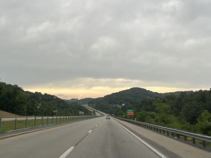 A highway where two lanes of southbound traffic and two lanes of northbound traffic are separated by a grass medium. The sky is turning yellow overhead, and a sign reads "Weigh Station" on the side of the road.
