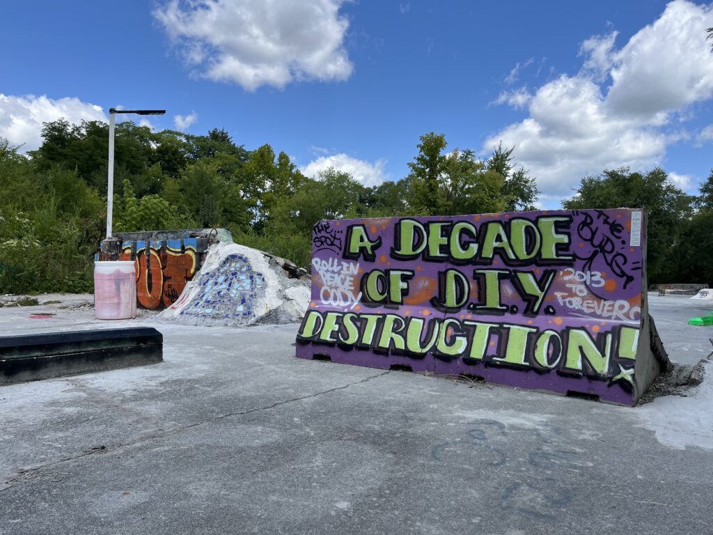 A concrete ramp on a flat lot reads the following words in purple and yellow spray painted text: "Decade of DIY Destruction!" Other ramps and skatepark features are visible in the background. Trees and a clear blue sky are visible in the distance.