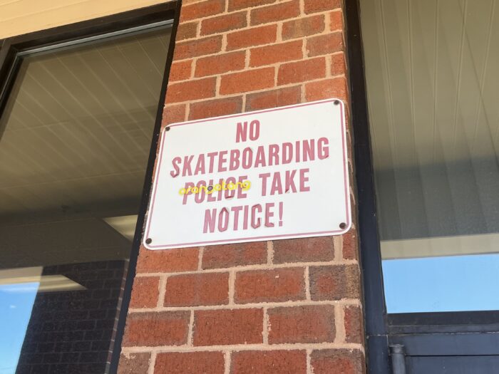 A red and white sign sits on the red-brick exterior of a building with tall glass pane windows. The sign reads, "NO Skateboarding. Police Take Notice!" A yellow sticker has been placed atop the word "police."