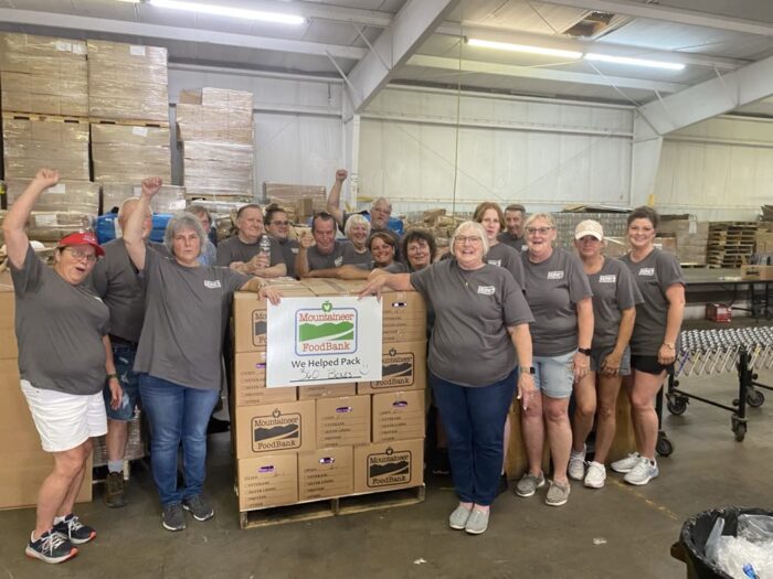 A group of people stand around a pile of boxes containing groceries. A sign on the boxes reads: "Mountaineer Food Bank."
