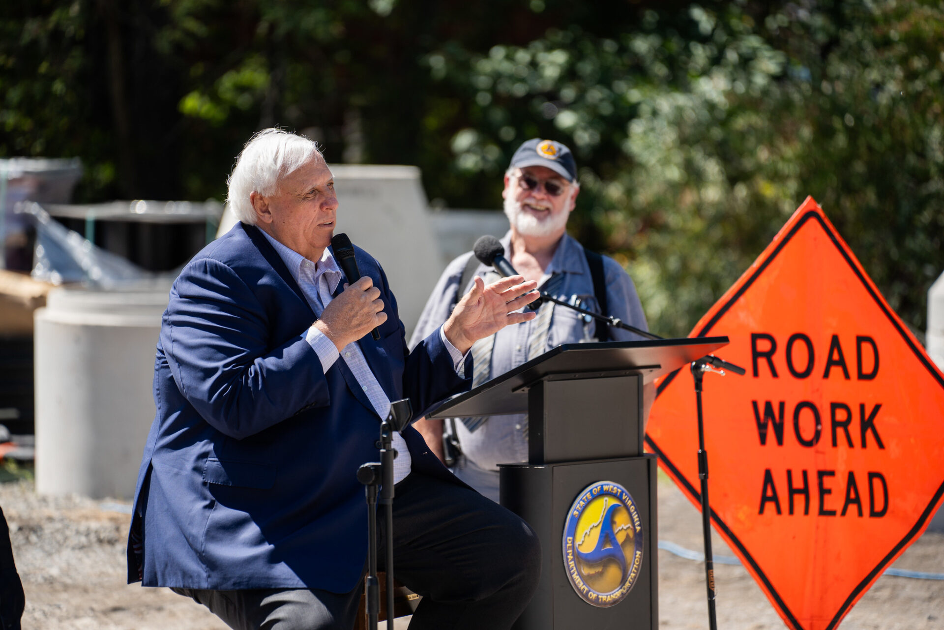 Man in dark suit sits on a stool in front of a road sign with a microphone