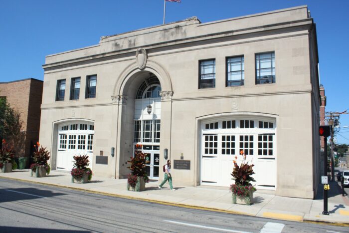 A light-colored stone building with large, white garage-style doors on either side of a multi-story entrance sits in front of a clear blue sky. In front of the building are potted plants and a man walks just to the right of the entrance. A brass sign is visible to the left of the man that reads "Morgantown Municipal Building"