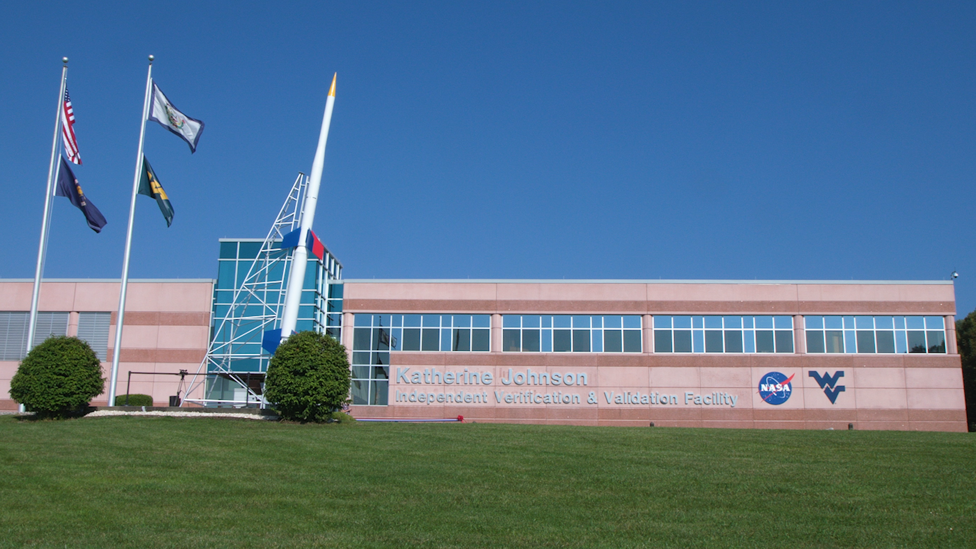 Flagpoles display four flags flapping in the breeze in front of a stone building of a reddish hue with blue-tinted glass windows running along the top edge. The building sits atop a green, grassy slope with a large model of a rocket in front.