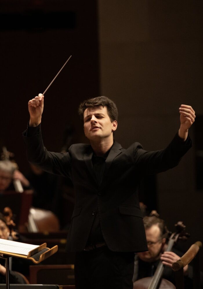 A man dressed in an all black suit raises his arms with his eyes closed. He holds a conductor's baton in his right hand, camera left. String players, including a celloist, can be seen behind him and out of focus. 