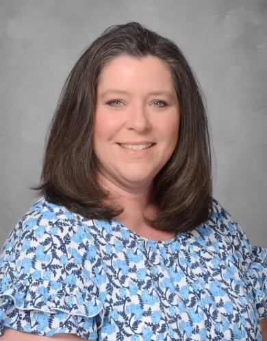 A portrait of a woman with shoulder-length brown hair in front of a light grey background. She wears a  light blue blouse with a white and dark blue pattern.