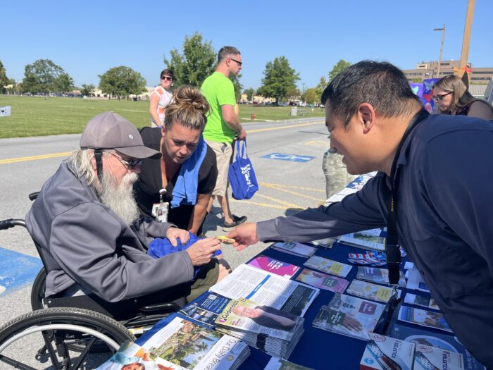 A man in a baseball hat wheelchair and a woman in a black t-shirt stand before a table with pamphlets and small items laid out atop a blue tablecloth. On the other side of the table, a man reaches over to hand something to the man.