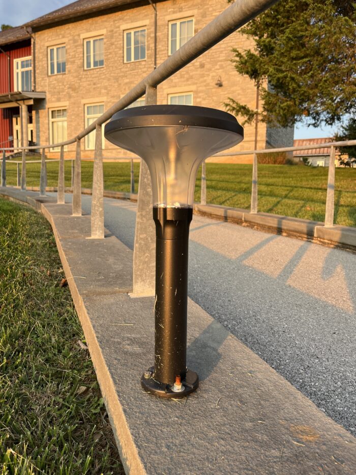 Along an asphalt pathway with a railing, a low-to-the-ground lighting fixture sits. It flares out at the top and is narrow toward the bottom and middle. It is black, except at the top it is translucent where the light is emitted. In the background, buildings and a lawn are visible.