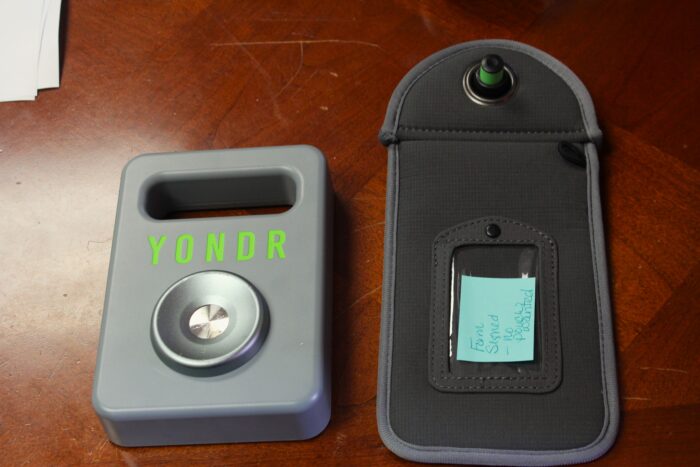 A grey rectangle with a handle at its top has the word "Yondr" stamped on it in a neon green color above a cupped metallic circle. To the right is a dark grey cloth bag with a plastic toggle up top and a small blue post-it with handwriting in the middle. Both items are on a dark wood surface, with the corner of a stack of papers in the top left corner.