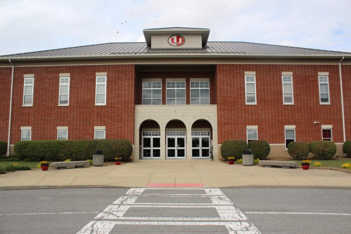 A brick two story facade of a school building is seen across a street. The center of the facade is recessed, with large windows on top and a three arch entryway on the ground floor.