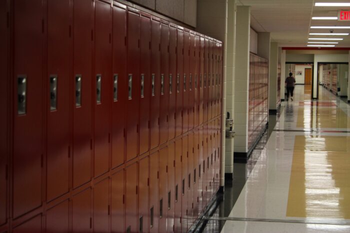 Red lockers extend from the left of frame into the center in a flourescent-lit school hallway. In the far distance, framed by a doorframe, an individual can be seen walking down the hall. The floor's linoleum showcases a black border, with yellow accents in the center and white in between.