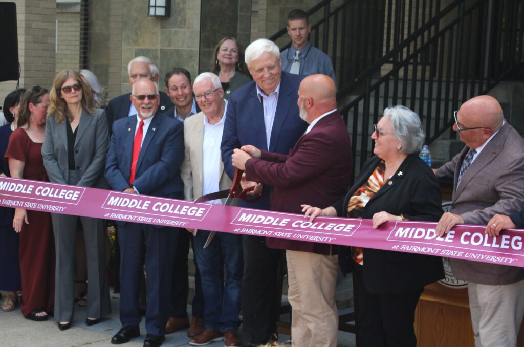 A row of people stand behind a dark red ribbon with the words "Middle College at Fairmont State University" printed in white on it. In the middle two men wearing suits hold large, ceremonial scissors poised to cut the ribbon. Behind them stairs with a black railing lead up and to the right of frame.