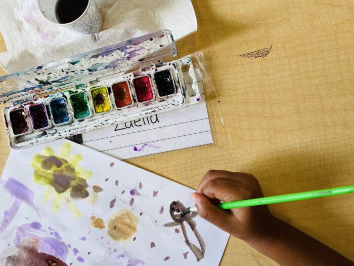 a palette of water colors sits on a table while a child's hand holds a brush and paints.