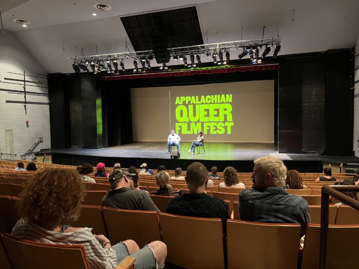 Two men sit on a stage in front of a large projector screen that reads "Appalachian Queer Film Fest" in large lettering. People sit somewhat spaced out down in the audience.