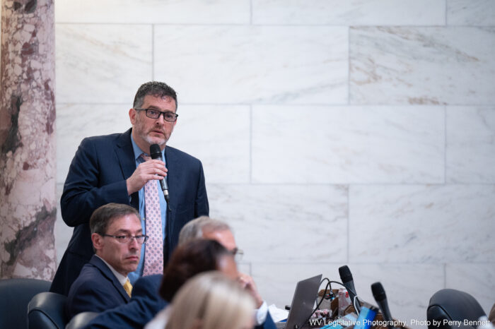 A man wearing glasses in a dark blue suit with a pink tie over a light blue shirt stands in front of a white marble wall holding a microphone to his mouth to speak. In front of him, another bespectacled man sits in a row of people that extends into the foreground.