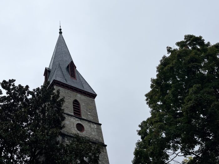 A stone brick church spire with a pointed top stands tall beside dark green trees.