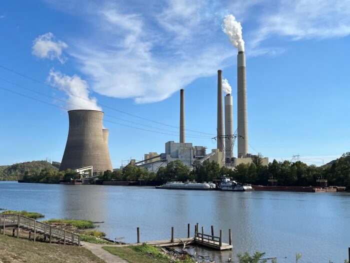 A power plant emits light plumes of steam through its stacks and a concrete cooling tower, with a placid river in the foreground and a clear sky behind it.