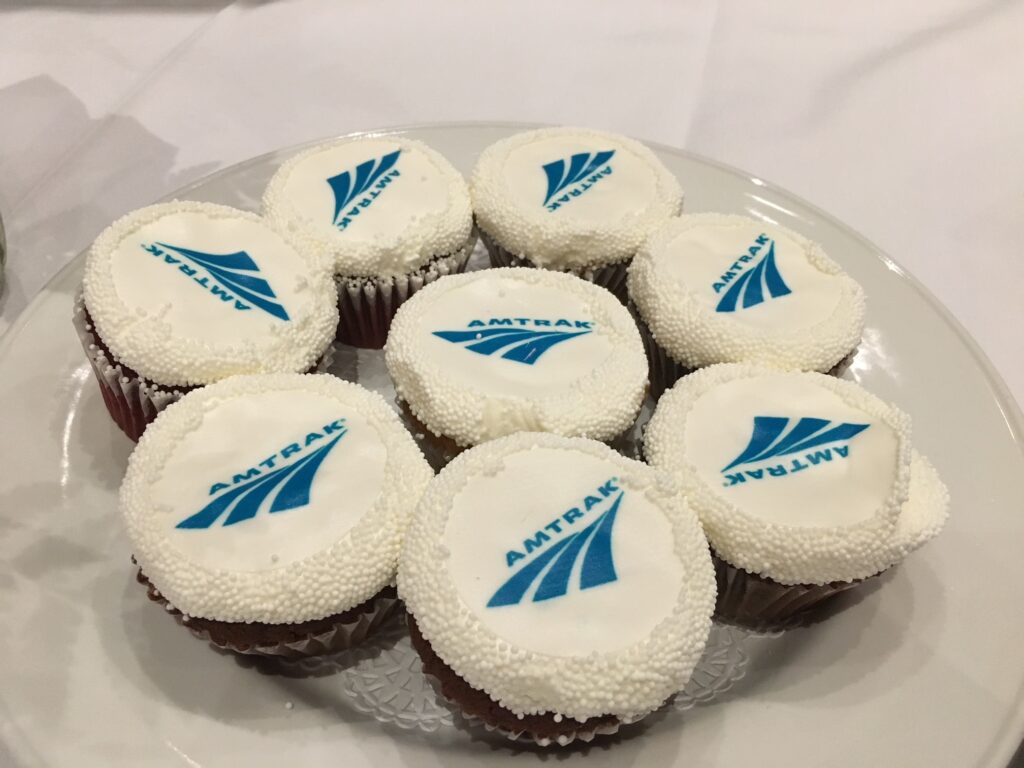 Cupcakes with white icing and a blue Amtrak logo are served on a plate sitting on a white tablecloth.