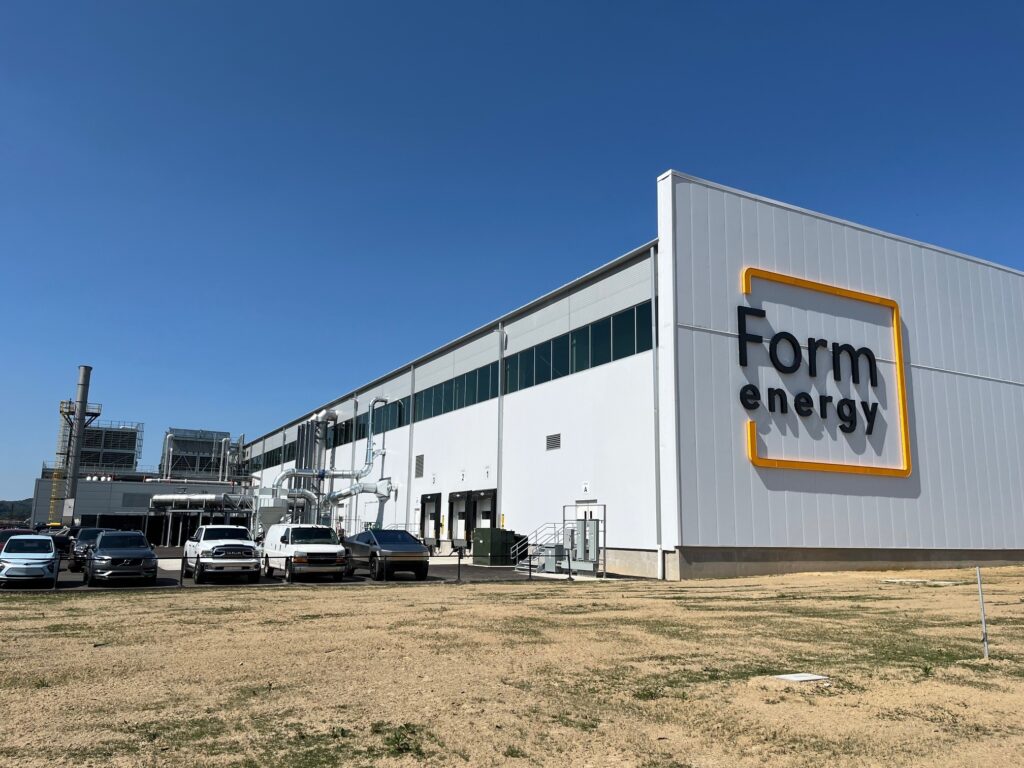 A white industrial building with cars parked next to it against a clear sky with dry, brown grass in the foreground.