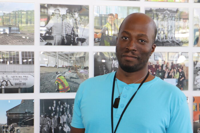 A man wearing a light blue shirt and a lanyard stands in front of a gallery of photos of workers at an old steel factory.