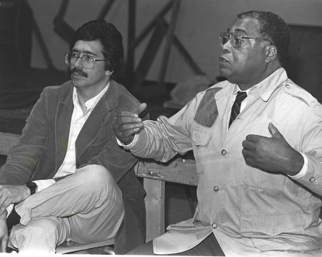 In a black and white image, a well-known actor expressively speaks to a group of students with the class instructor on his right. Both men are dressed of the period.