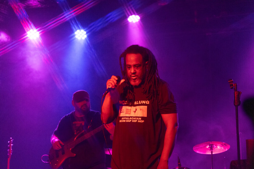 An African American man with long hair holds a microphone and stands on stage in front of a band.