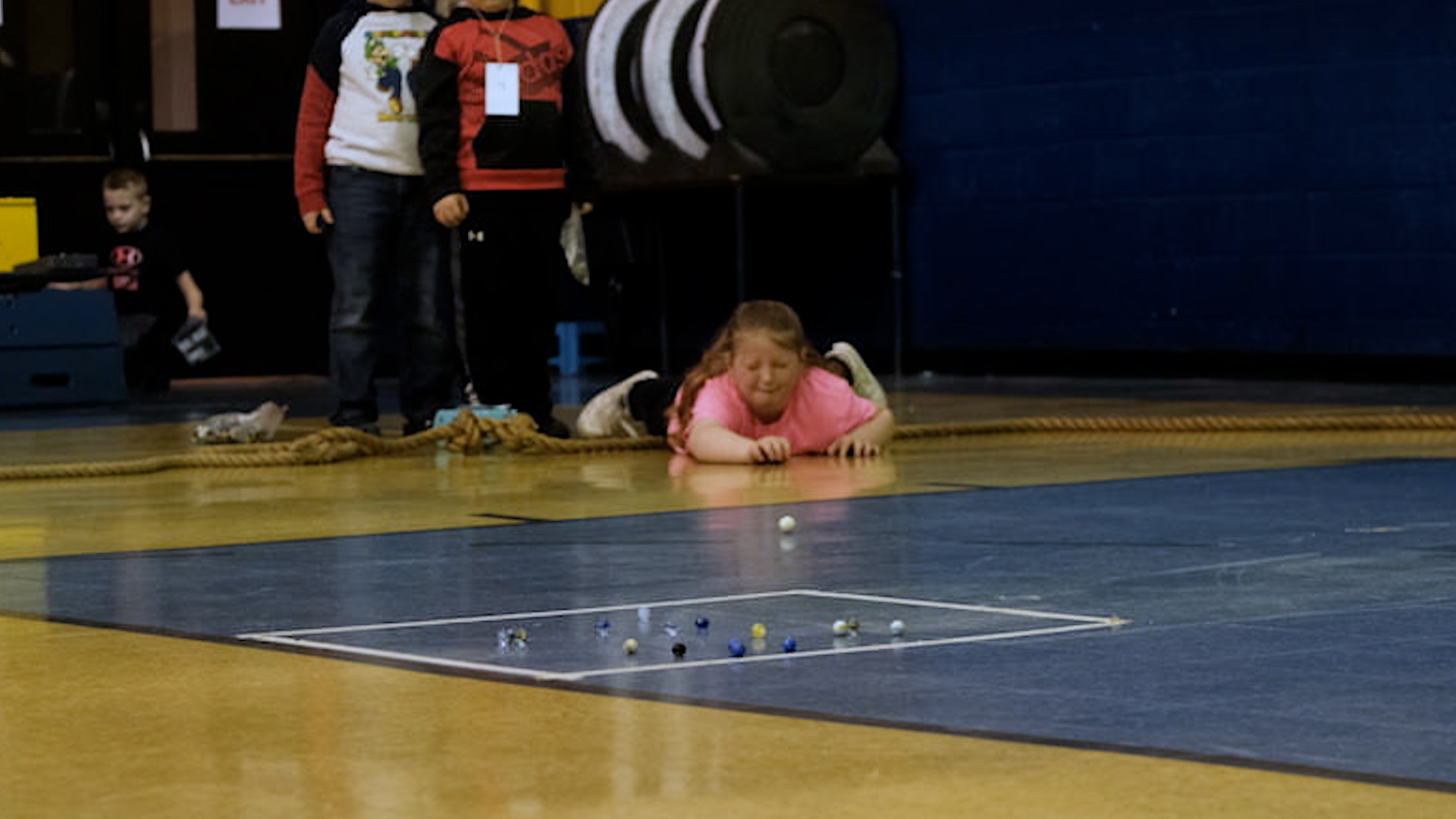 A girl lays across a gym floor and shoots a white marble toward a group of other marbles.