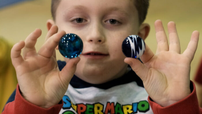 A young boy holds up a clear, blue marble in one hand and a blue and white marble in the other.