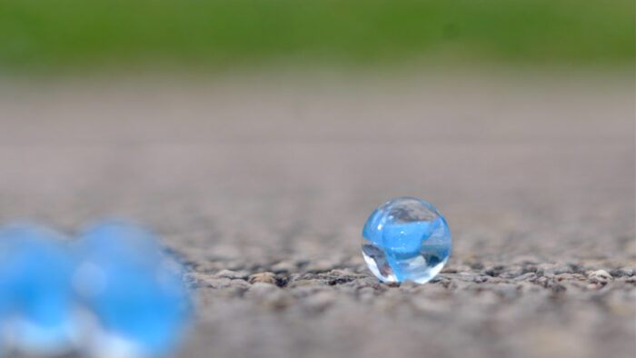 A single blue marble in clear focus on pavement with several other similar marbles to the side and blurry.