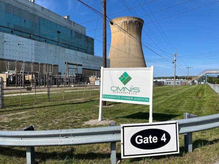 A sign for a power plant next to a state highway with a concrete cooling tower behind it against a clear sky.