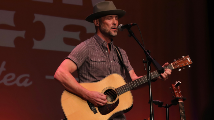 A man wearing a hat plays a guitar with his eyes shut while he sings into a mic.