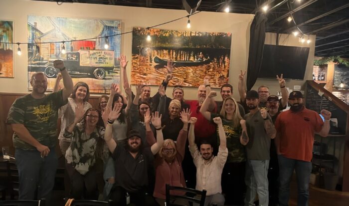 A crowd of people stand together in a restaurant, facing the camera. They are smiling and throwing their arms in the air in celebration.