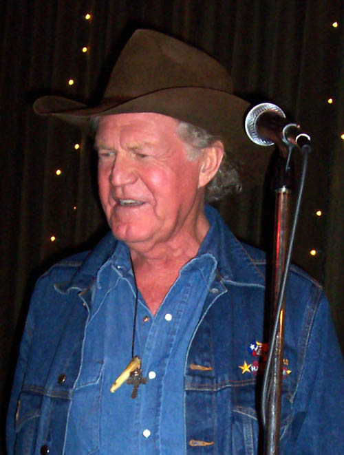 A man in a brown felt hat wears a dark blue denim jacket over a lighter blue denim shirt with pearl buttons. He stands just behind a wired microphone in a stand and in front of a brown curtain with fairy lights strung over it. The man wears a necklace with a cross pendant, as well as a bone pendant. 