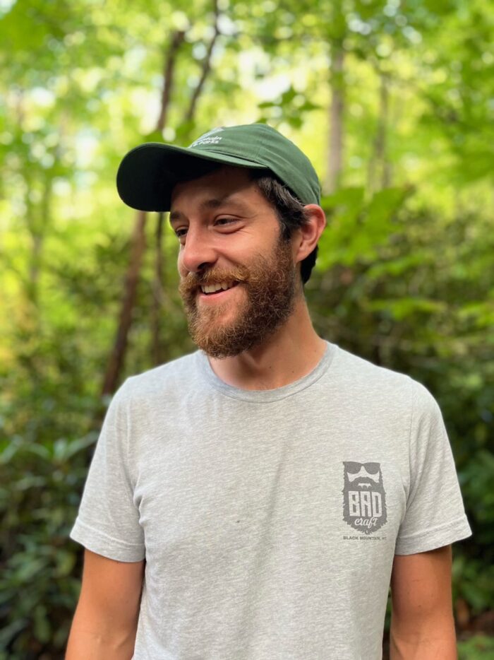 A young, bearded man in a green baseball cap and a t-shirt stands in the woods.